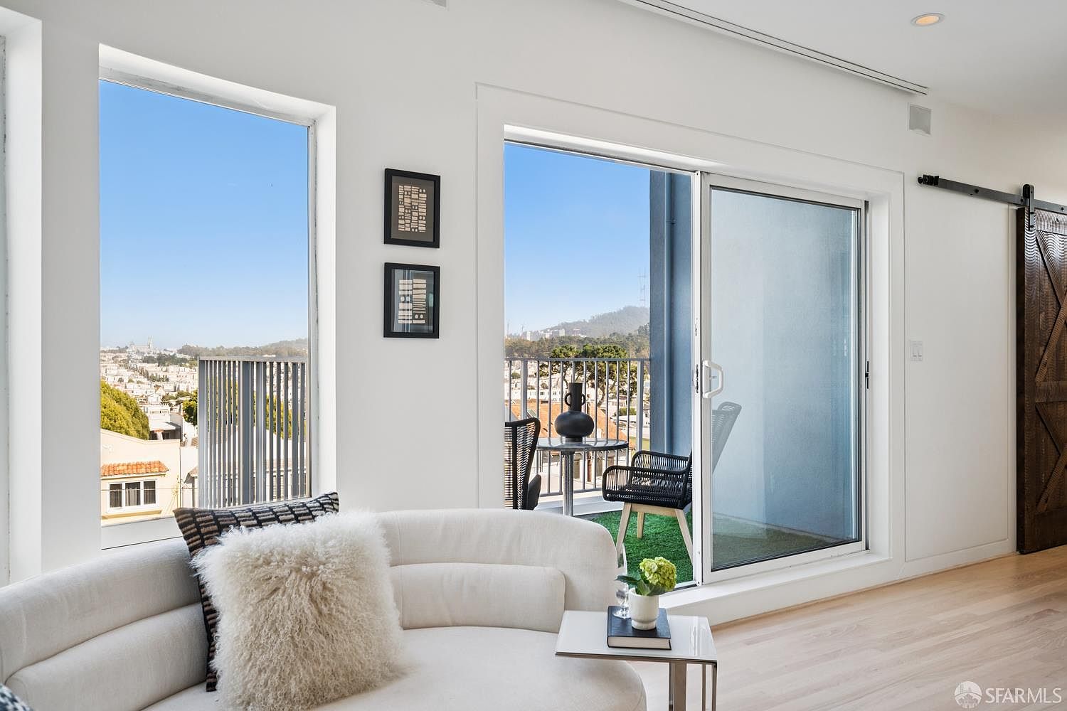 This bright, modern living room features a curved white sofa adorned with a textured throw pillow, positioned near a large window and a sliding glass door that opens onto a private balcony. The space is characterized by light wood flooring, crisp white walls, and a minimalist aesthetic, with a small side table holding a decorative plant and books. The balcony offers an outdoor seating area with a view of the surrounding neighborhood and hills, creating a seamless transition between indoor and outdoor living.