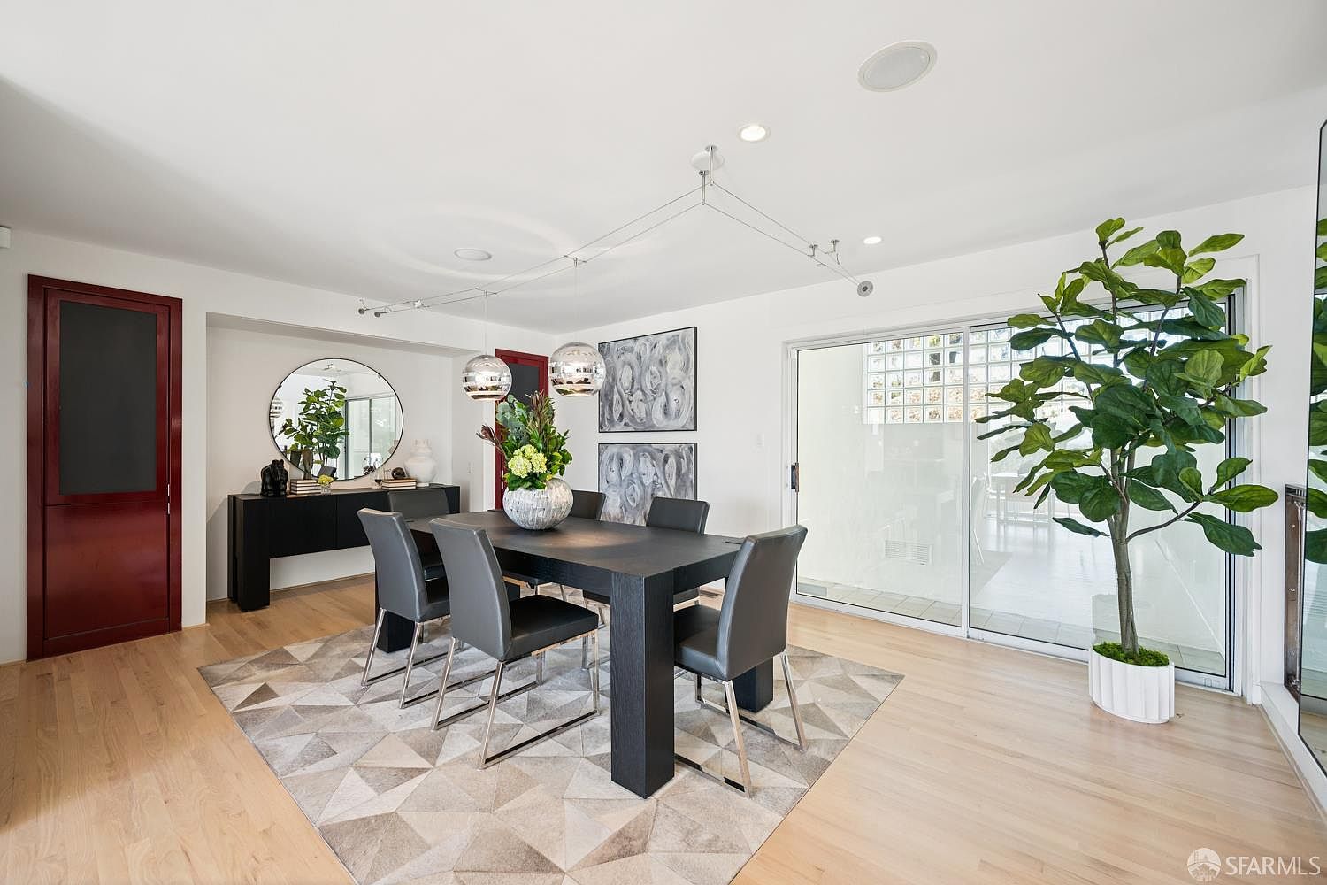 This contemporary dining room features a dark wood table surrounded by six grey upholstered chairs, set atop a geometric patterned area rug. A sleek black console table sits against the wall beneath a large circular mirror, while a tall potted plant adds a touch of greenery near the sliding glass doors. The space is illuminated by modern pendant lighting and recessed ceiling lights, creating a bright and sophisticated atmosphere.