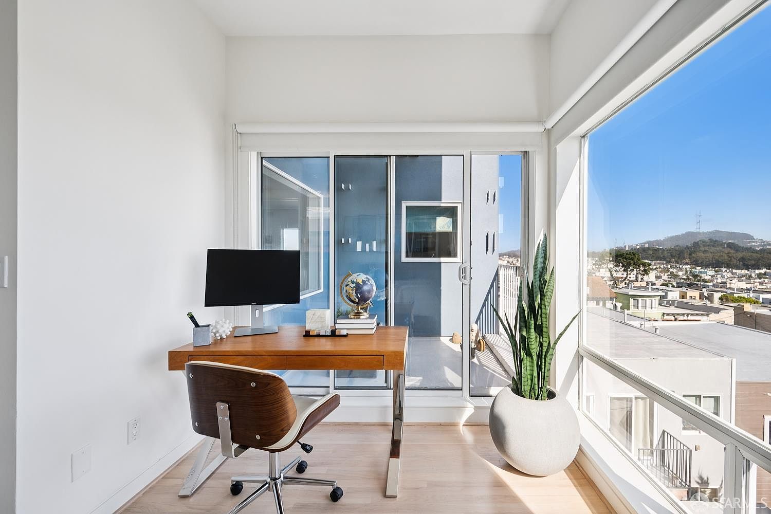 This bright home office features a sleek wooden desk positioned to face a large window, offering an expansive view of the surrounding neighborhood and hills. The room is minimalist in design, accented by a modern mid-century style chair, a potted snake plant, and a globe on the desk. Large glass sliding doors lead out to a private balcony, filling the space with natural light and creating an airy, productive atmosphere.