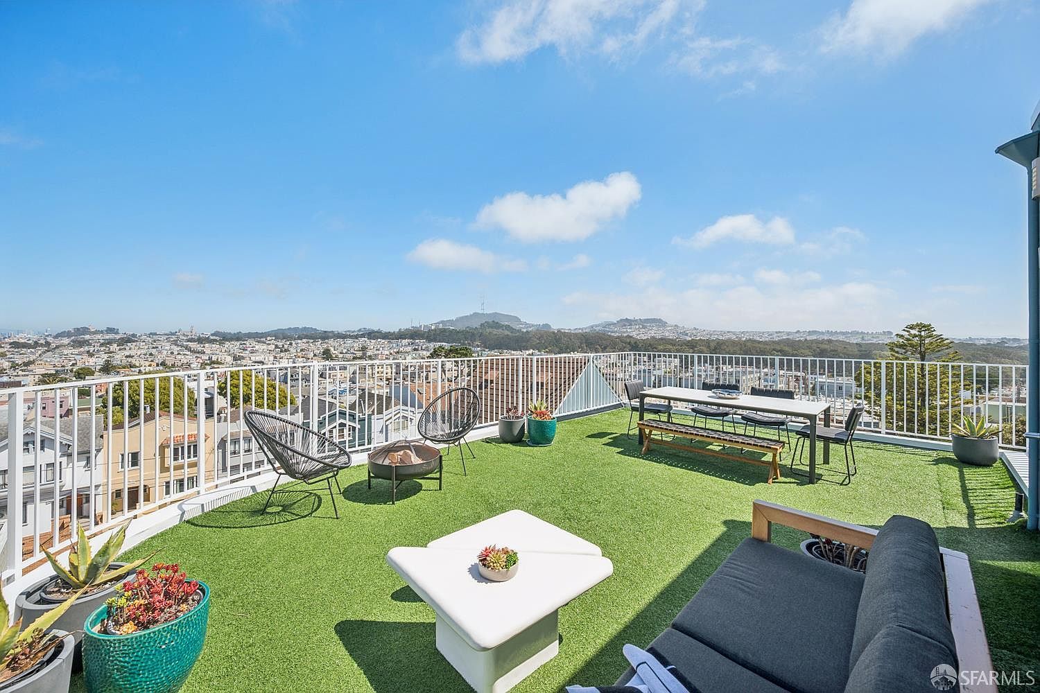 This expansive rooftop deck offers a panoramic view of the surrounding cityscape under a bright, clear sky. The space is furnished with a modern outdoor sofa, a dining set, and casual lounge chairs arranged on artificial turf, creating an inviting area for relaxation or entertaining. A small fire pit and several potted plants add warmth and greenery to this elevated urban retreat.
