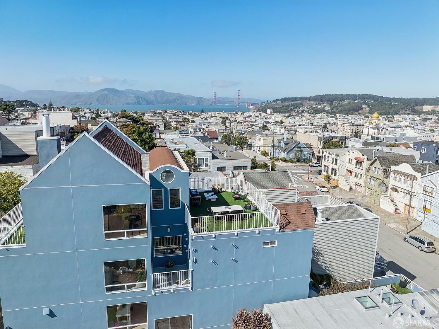 This elevated aerial perspective captures a blue-sided multi-story residence featuring a prominent rooftop deck with artificial turf and outdoor seating. The property is situated in a dense urban neighborhood, offering expansive views of the city skyline and the Golden Gate Bridge in the distance. The composition highlights the home's architectural profile, including its varied rooflines and multiple levels of windows, set against a clear blue sky.