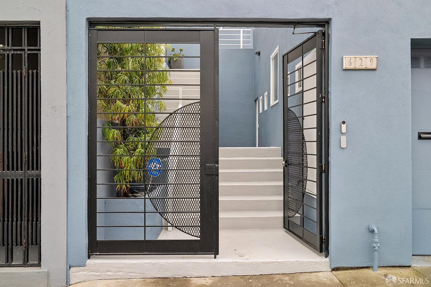 This entryway features a modern, light blue facade with a prominent black metal security gate that is partially open. Beyond the gate, a set of light-colored stairs leads upward, framed by a small, lush green plant in a planter. The address '629' is clearly displayed on a rectangular plaque to the right of the entrance, which also includes a doorbell and a wall-mounted mailbox.