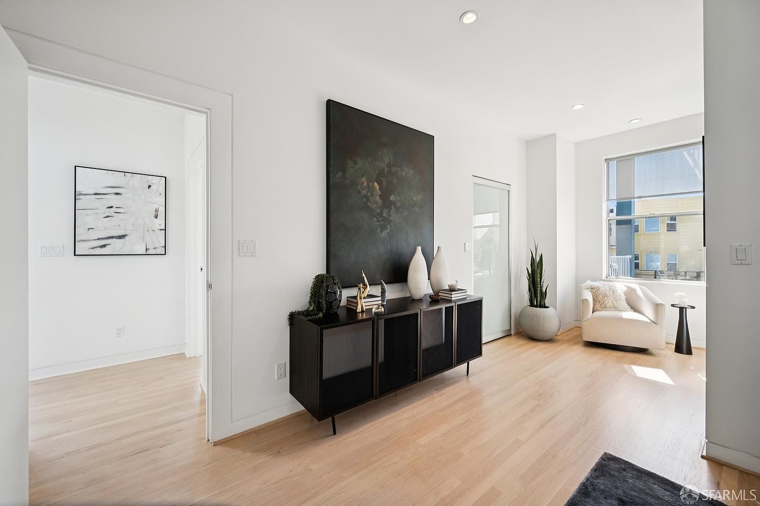 This bright and modern living space features light wood flooring and crisp white walls, creating an airy and open atmosphere. A sleek black sideboard sits against the wall, topped with decorative vases and a large, dark abstract painting, while a comfortable cream-colored armchair is positioned near a large window. The perspective captures a clean, minimalist aesthetic with a doorway leading to an adjacent room, emphasizing the home's contemporary design.
