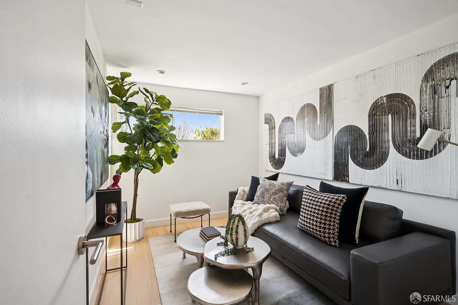 This modern living room features a sleek black leather sofa adorned with patterned throw pillows and a soft white throw blanket. A large, abstract black-and-white wall art piece serves as a bold focal point, complemented by a tiered metal coffee table and a tall potted fiddle leaf fig tree. The space is brightened by a window and light wood flooring, creating a sophisticated and contemporary atmosphere.
