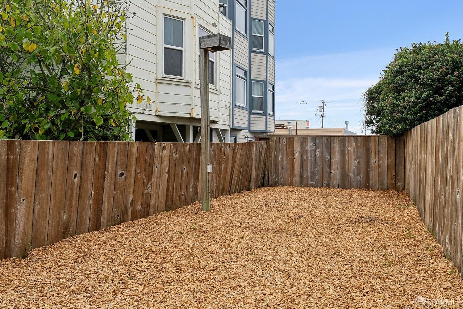 This backyard space features a ground cover of wood chips enclosed by a tall, weathered wood privacy fence. A single wooden post stands in the center of the yard, and the rear of a multi-story residential building is visible in the background. The area provides a private, low-maintenance outdoor space with a clear view of the adjacent structure.