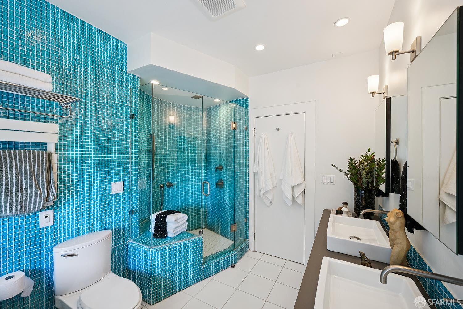 This vibrant primary bathroom features striking turquoise mosaic tile work that covers the shower enclosure and the adjacent wall. The space includes a double vanity with white rectangular sinks, a glass-enclosed walk-in shower with a built-in bench, and a white toilet. The bright, modern aesthetic is enhanced by recessed lighting and a large mirror, creating a clean and energetic atmosphere.