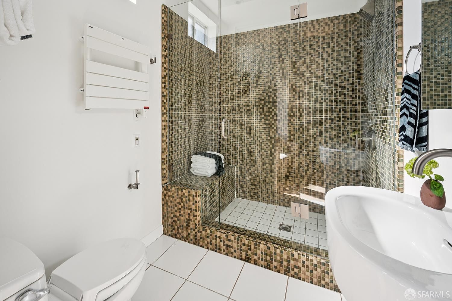 This modern bathroom features a walk-in shower enclosed by a glass panel, showcasing intricate brown and gold mosaic tile work on the walls and a built-in bench. The space includes a white pedestal sink, a wall-mounted towel warmer, and a toilet, all set against clean white floor tiles. The overall aesthetic is contemporary and functional, with a focus on textured surfaces and efficient use of space.