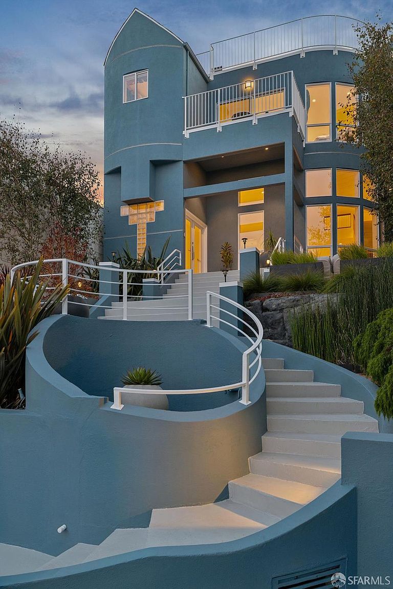 This striking contemporary home features a bold, blue-toned facade with a unique, multi-level architectural design. A dramatic, curved staircase with white railings leads up to the main entrance, creating a grand and inviting approach. The exterior is accented by large windows, a glass block feature, and tiered landscaping, all captured from a low-angle perspective that emphasizes the home's height and modern aesthetic.