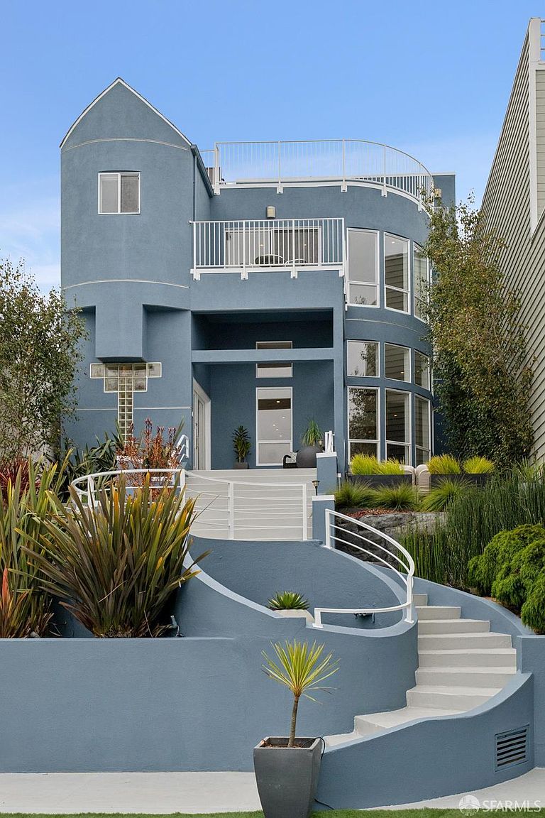 This striking contemporary home features a unique blue stucco facade with a prominent curved architectural design and a multi-level balcony structure. A sweeping, curved staircase with white railings leads up to the main entrance, complemented by tiered landscaping and ornamental grasses. The perspective is a low-angle, eye-level shot that emphasizes the home's verticality and modern, sculptural aesthetic.