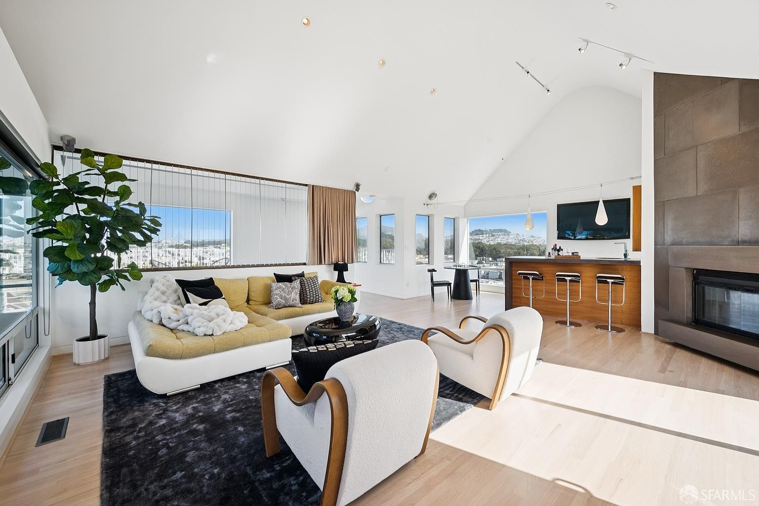 This bright and airy living room features a vaulted ceiling and an open-concept layout that flows into a bar area. A large, light-colored sectional sofa and two curved accent chairs are arranged on a dark area rug, creating a comfortable seating area near a tall, stone-clad chimney breast. Large windows provide expansive views of the surrounding landscape, while light wood flooring and minimalist decor contribute to a modern, sophisticated aesthetic.