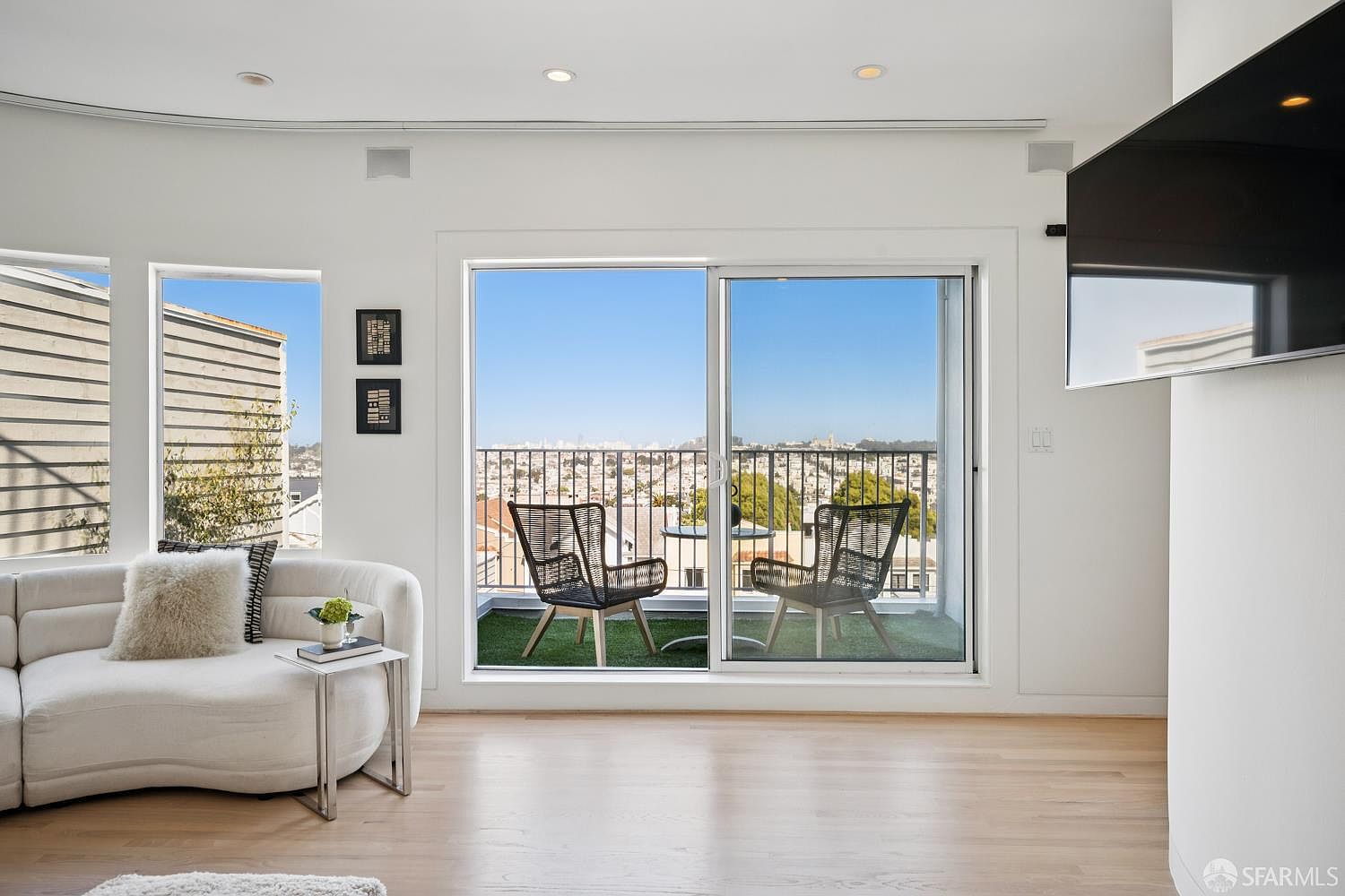 This bright and airy living room features light wood flooring and a curved, cream-colored sofa accented with a textured throw pillow. A large sliding glass door provides access to a private balcony with outdoor seating and a panoramic city view. The minimalist aesthetic is complemented by recessed lighting and a wall-mounted television, creating a clean and inviting atmosphere.