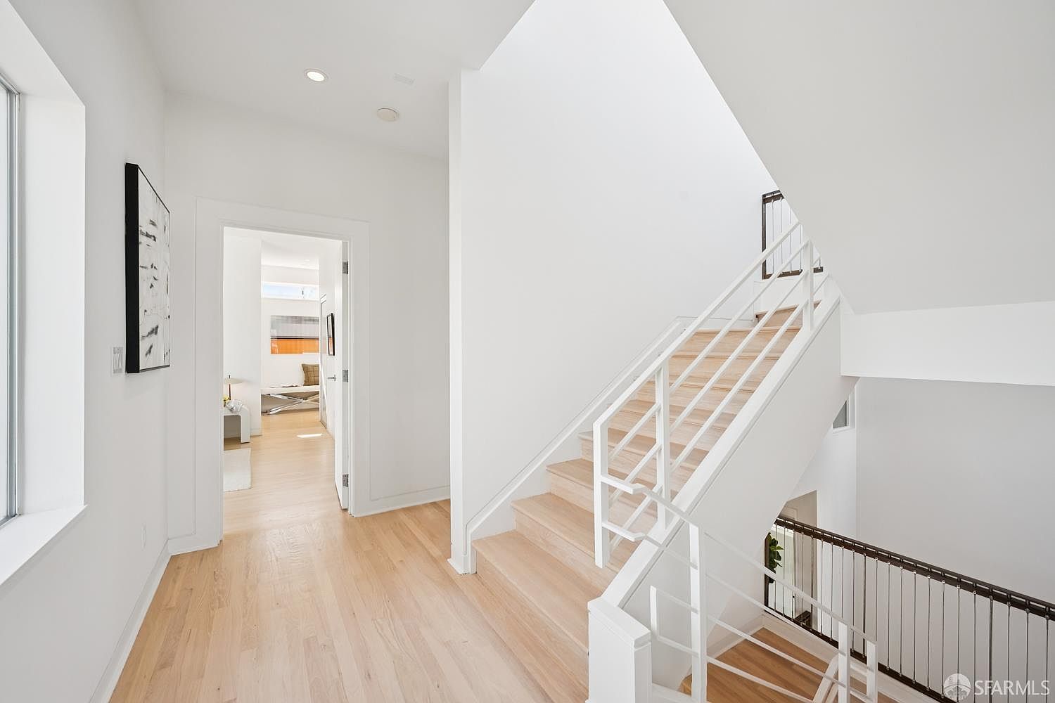 This bright, minimalist hallway features light wood flooring and crisp white walls, creating an airy and modern atmosphere. A staircase with white railings and light wood treads leads both upward and downward, while an open doorway provides a glimpse into a sunlit room beyond. The clean lines and neutral palette emphasize a contemporary architectural style.