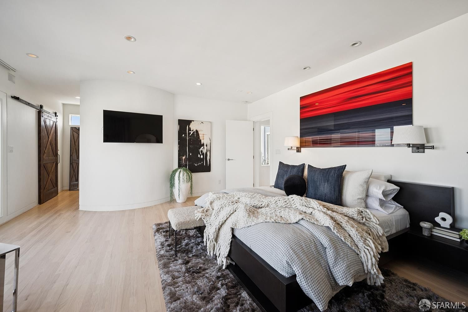 This spacious primary bedroom features light wood flooring, a plush gray area rug, and a modern platform bed dressed in textured linens. A striking red and black abstract art piece hangs above the headboard, complemented by a curved wall section that houses a wall-mounted television. The room is accented by barn-style sliding doors and minimalist wall sconces, creating a sophisticated and contemporary atmosphere.