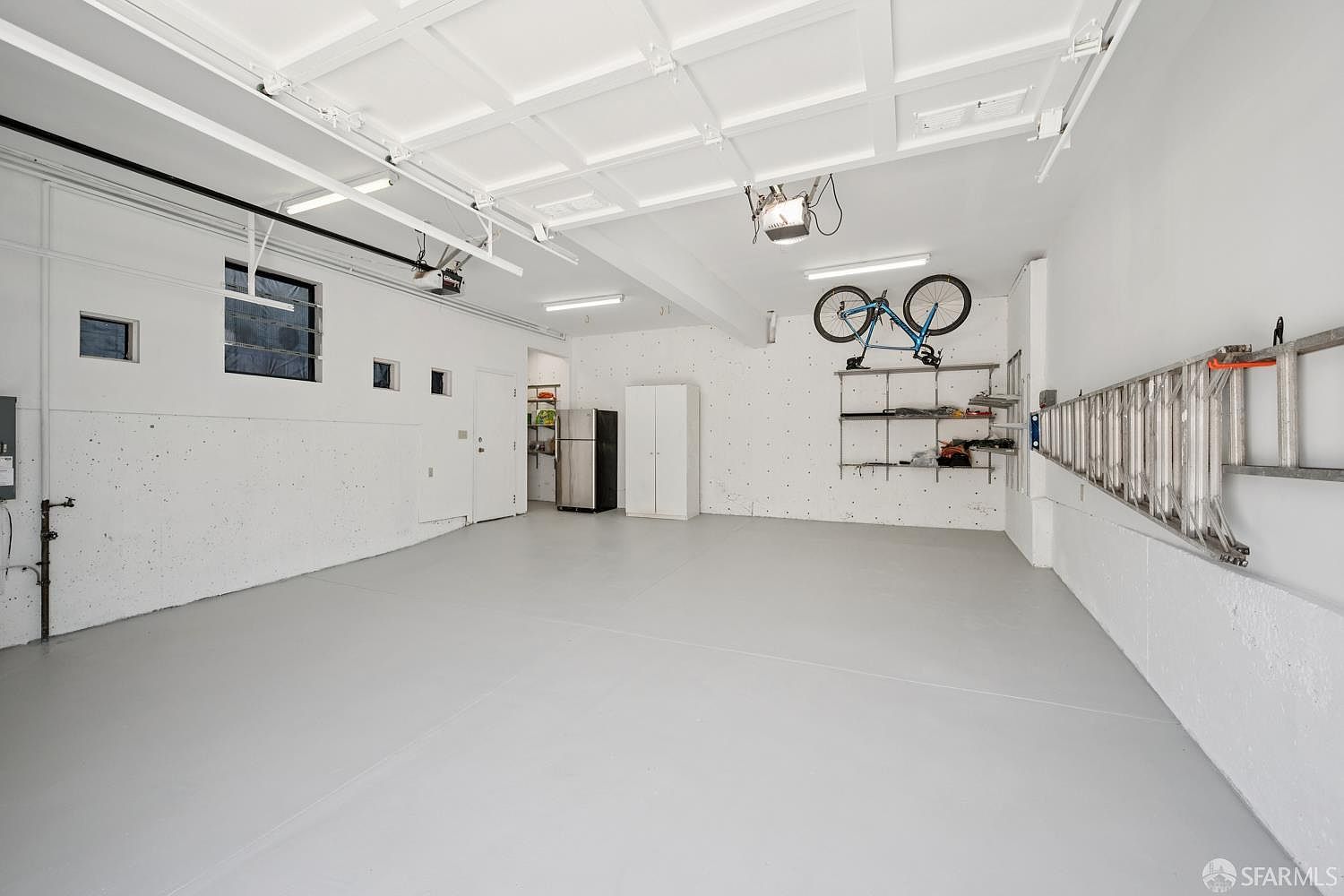 This spacious, well-lit garage features a clean, minimalist aesthetic with white walls and a light-colored epoxy floor. The area includes a wall-mounted storage rack holding a bicycle, a metal shelving unit, and a refrigerator, providing functional utility space. The perspective is wide-angle, capturing the depth of the garage and the overhead door mechanism.