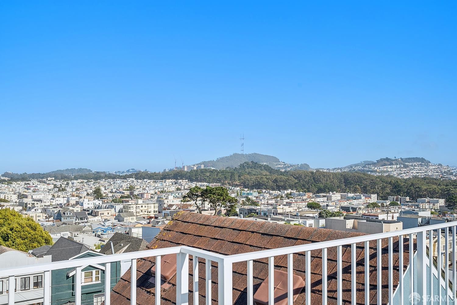 This image captures a wide, elevated view from a balcony, showcasing a dense urban landscape under a clear blue sky. In the foreground, a white metal railing frames the scene, overlooking a shingled roof and the sprawling city neighborhood below. The perspective emphasizes the expansive horizon and the hilly terrain in the distance, providing a sense of height and openness.