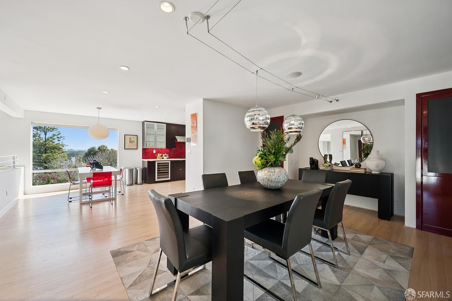 This open-concept dining area features a dark wood table surrounded by modern chairs, set atop a geometric patterned rug. The space is illuminated by contemporary pendant lights and recessed lighting, with a large circular mirror reflecting the room's depth. In the background, a kitchen with red tile backsplash and dark cabinetry is visible, while a large window provides a scenic view of the outdoors.