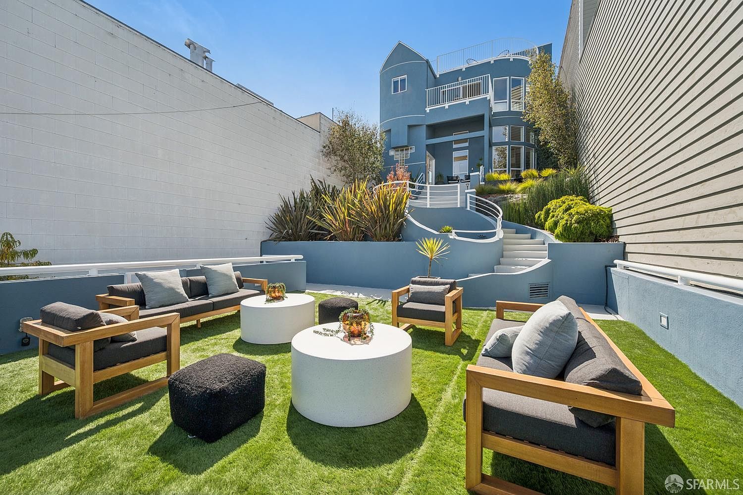 This backyard features a tiered layout with a synthetic turf lawn furnished with modern outdoor seating, including two sofas, two armchairs, and two round coffee tables. A curved staircase with white railings leads up to the blue-painted multi-story house, which showcases a contemporary architectural style with balconies and large windows. The space is enclosed by high walls, creating a private and well-maintained outdoor living area.