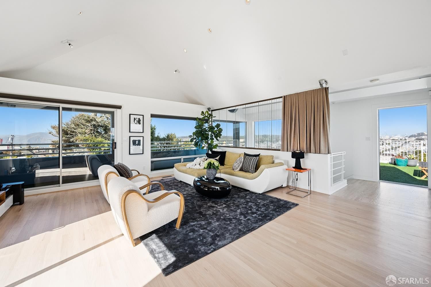This bright and airy living room features a vaulted ceiling and light wood flooring, creating an open and inviting atmosphere. The space is furnished with a modern white sectional sofa, two curved-back armchairs, and a dark, textured area rug, all centered around a sleek black coffee table. Large sliding glass doors and windows provide expansive views of the outdoors, while a curtained partition adds a touch of texture to the minimalist design.