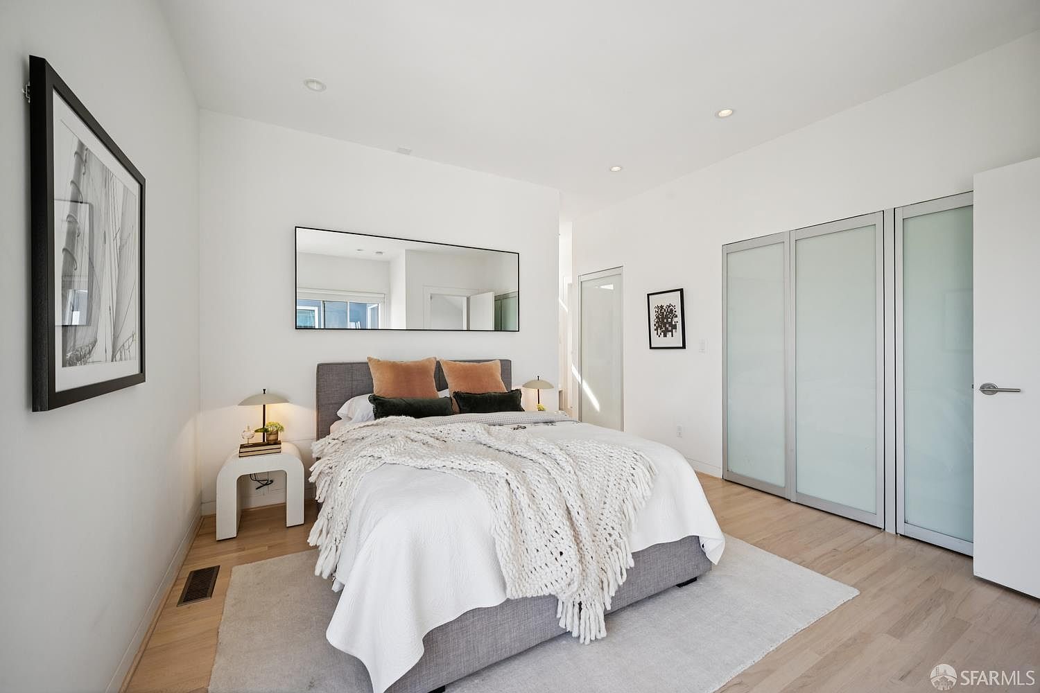 This modern primary bedroom features a neutral color palette with light wood flooring and crisp white walls. A gray upholstered bed is centered against the wall, accented by textured throw pillows and a chunky knit throw blanket, while a long, rectangular mirror hangs above the headboard to enhance the sense of space. To the right, a set of sliding glass-panel closet doors provides a sleek storage solution, and a framed piece of art adds a touch of personality to the minimalist design.