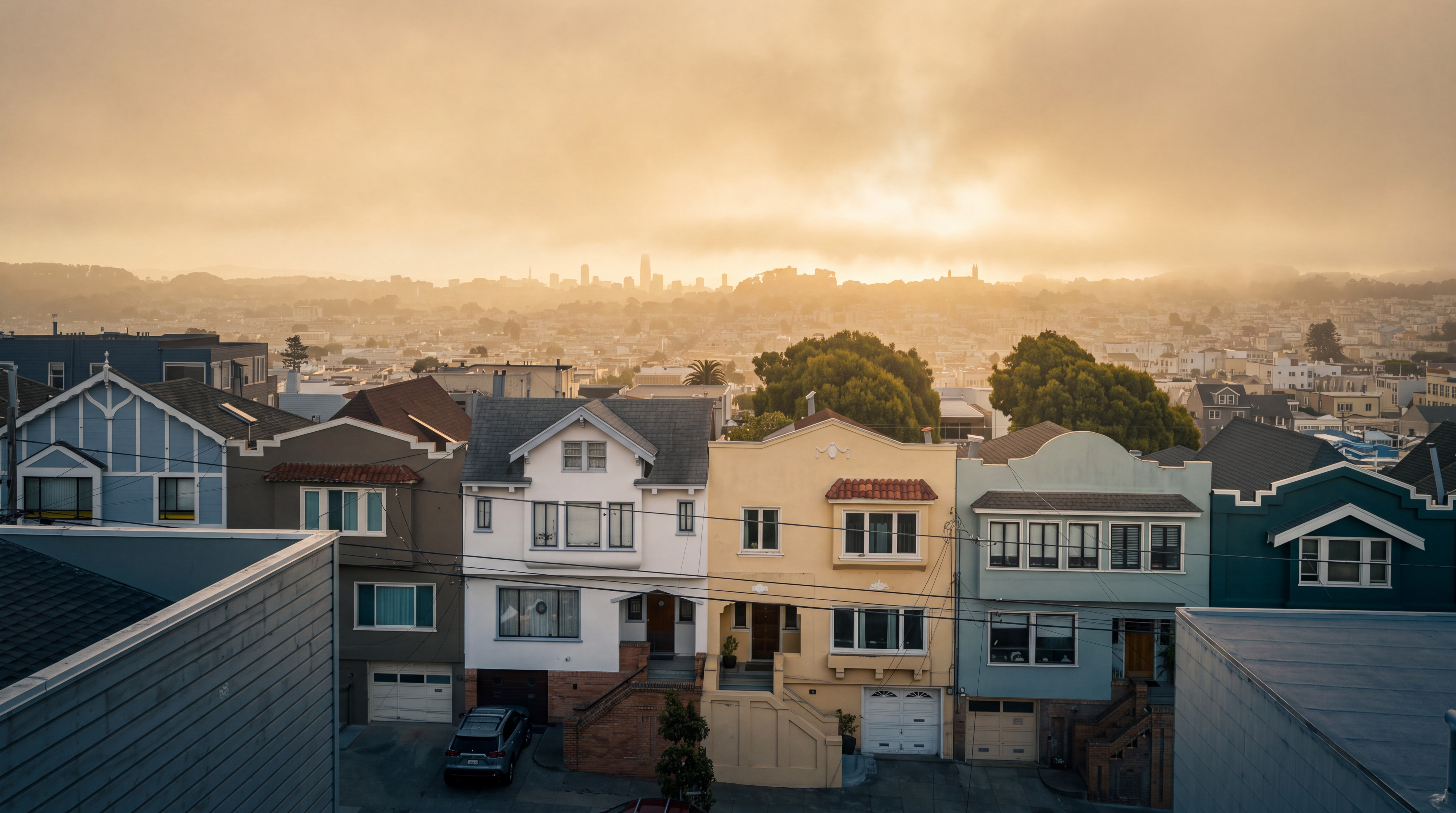 This elevated perspective captures a row of charming, multi-story residential homes in a dense urban setting, likely San Francisco. The architecture features a mix of traditional styles with varied pastel and neutral facades, bay windows, and pitched rooflines. Beyond the immediate streetscape, the image offers a sweeping, panoramic view of the sprawling city skyline under a clear blue sky.