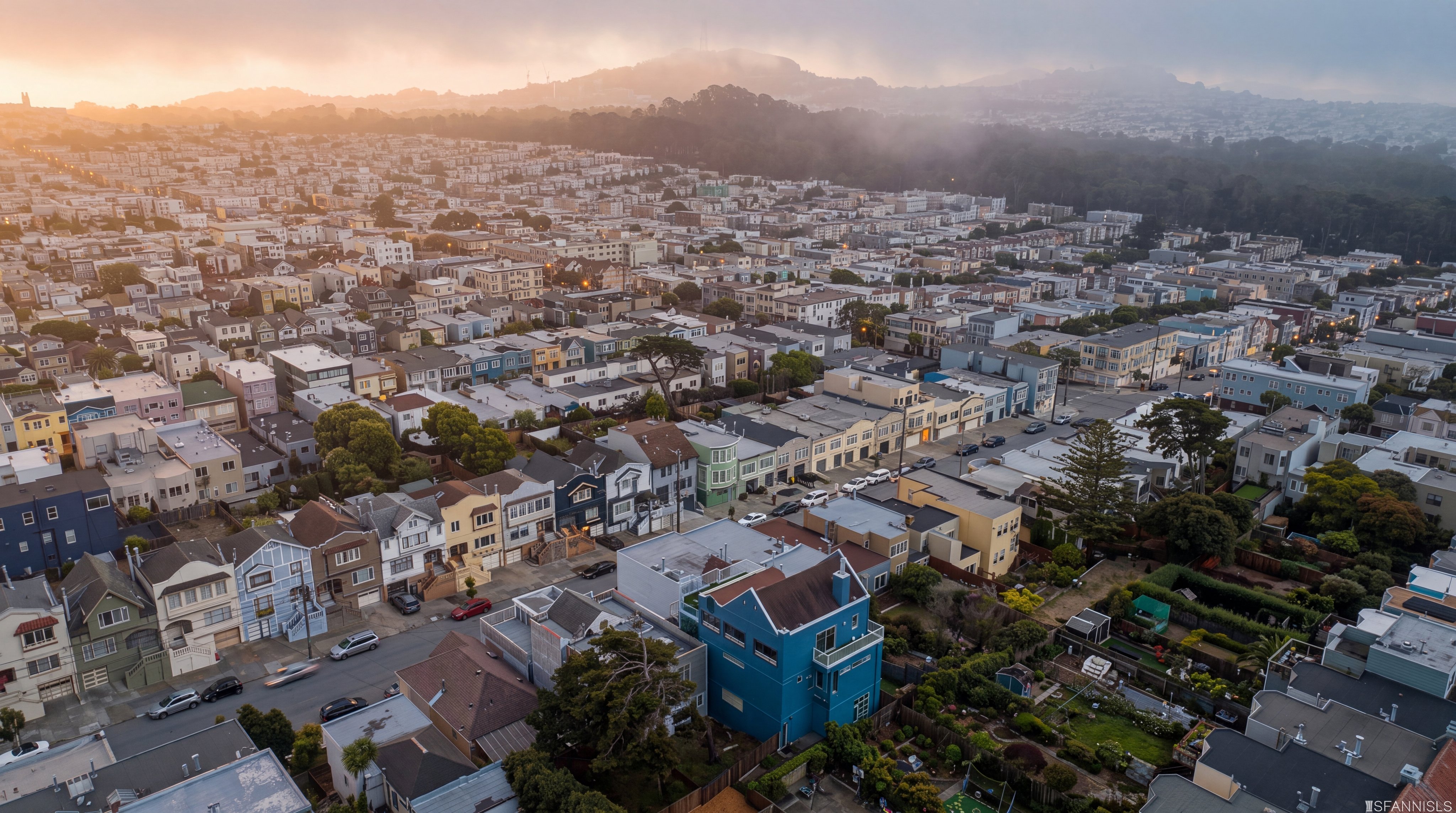 This high-angle aerial view captures a dense urban neighborhood in San Francisco, characterized by a mix of colorful multi-story residential buildings and lush green trees. A prominent blue house stands out in the foreground, featuring a backyard garden and a deck, while the background reveals rolling hills and a distant communications tower under a clear blue sky. The perspective provides a comprehensive look at the neighborhood's layout, street patterns, and proximity to natural landscapes.