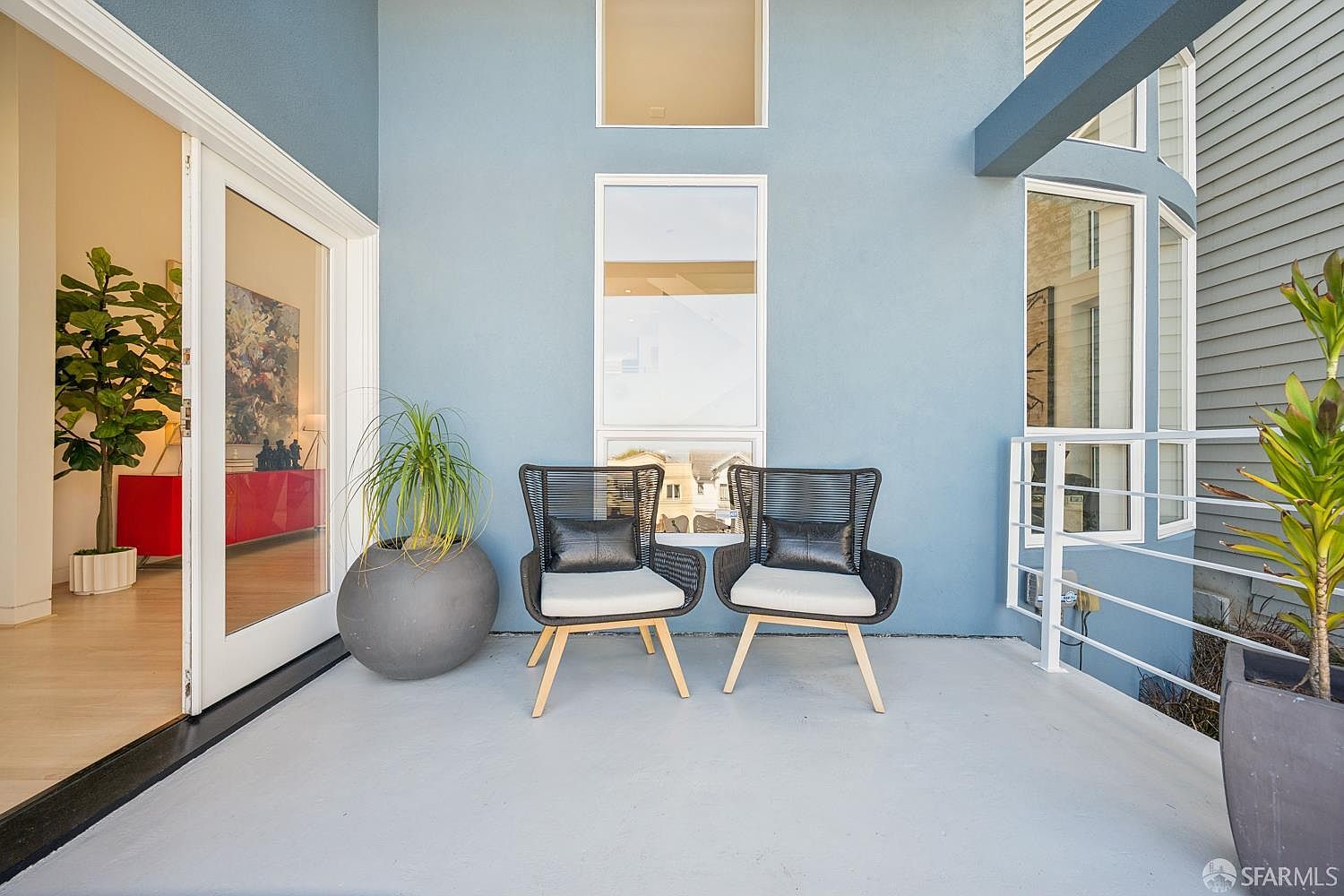 This modern balcony features a vibrant blue wall accented by two contemporary black woven chairs with light cushions and a large, spherical planter. The space is framed by a white metal railing and offers a glimpse into the interior living area through a glass door, where a bright red console table is visible. The perspective is eye-level, capturing a clean, inviting outdoor seating area that blends indoor and outdoor living.
