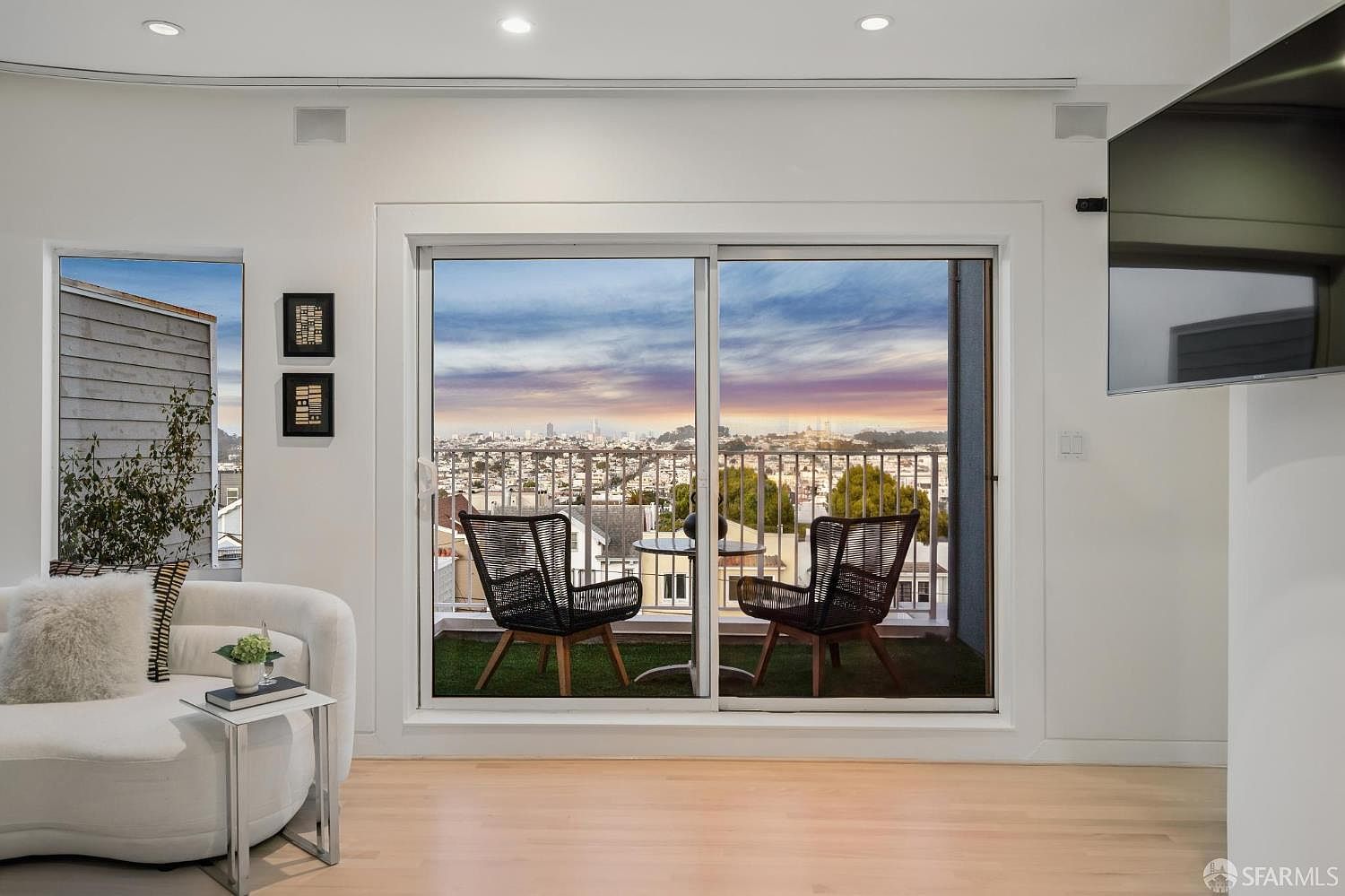 This bright living room features light wood flooring and a curved white sofa, creating a clean and modern aesthetic. A large sliding glass door provides access to a balcony with outdoor seating and a panoramic city view. The space is accented by two framed pieces of art on the wall and recessed ceiling lighting, offering a sophisticated and airy atmosphere.
