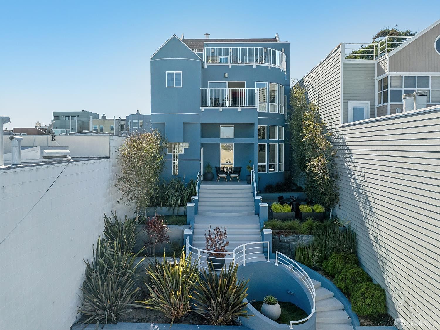 This rear view of a multi-level blue home showcases a tiered outdoor living space with a central staircase leading up to a patio area. The property features a curved, sunken seating area at the base, surrounded by manicured landscaping and mature plants. Large windows and multiple balconies provide a clear connection between the indoor and outdoor spaces, creating a modern and inviting atmosphere.
