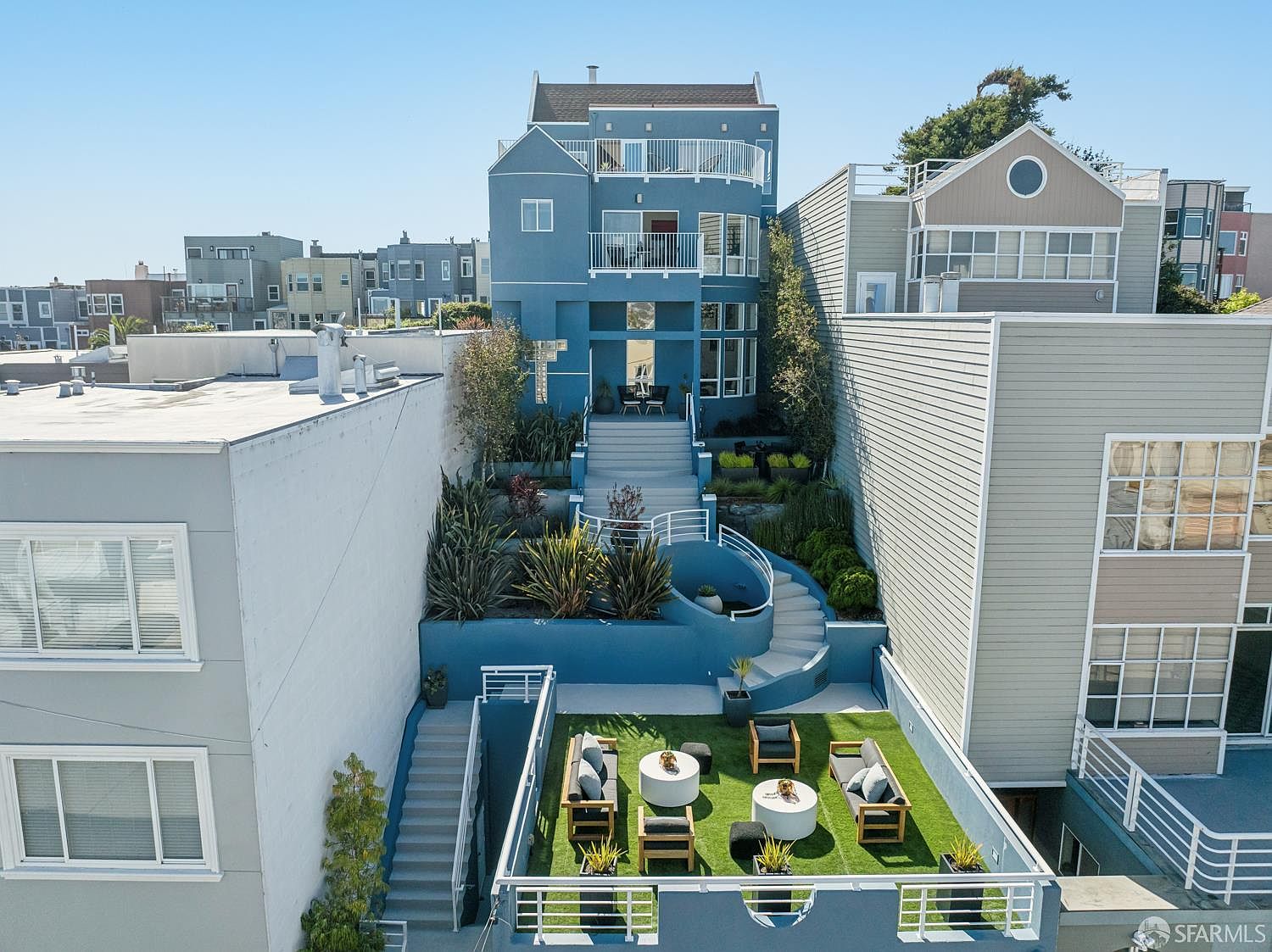 This aerial perspective captures a multi-level blue residential property with a tiered backyard design. The outdoor space features a synthetic turf patio area with lounge furniture, leading up a series of stairs through landscaped garden beds to the main house structure. The home showcases multiple balconies and large windows, reflecting a contemporary architectural style set within a dense urban neighborhood.