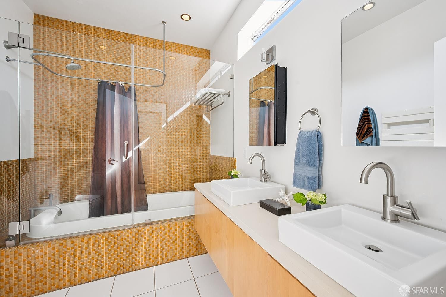 This modern bathroom features a double vanity with light wood cabinetry and rectangular vessel sinks, complemented by a large mirror and a blue towel hanging on a ring. A glass-enclosed shower and tub combination is lined with vibrant orange mosaic tiles, creating a bold focal point. The space is bright and clean, with white floor tiles and a high, narrow window that allows natural light to filter into the room.