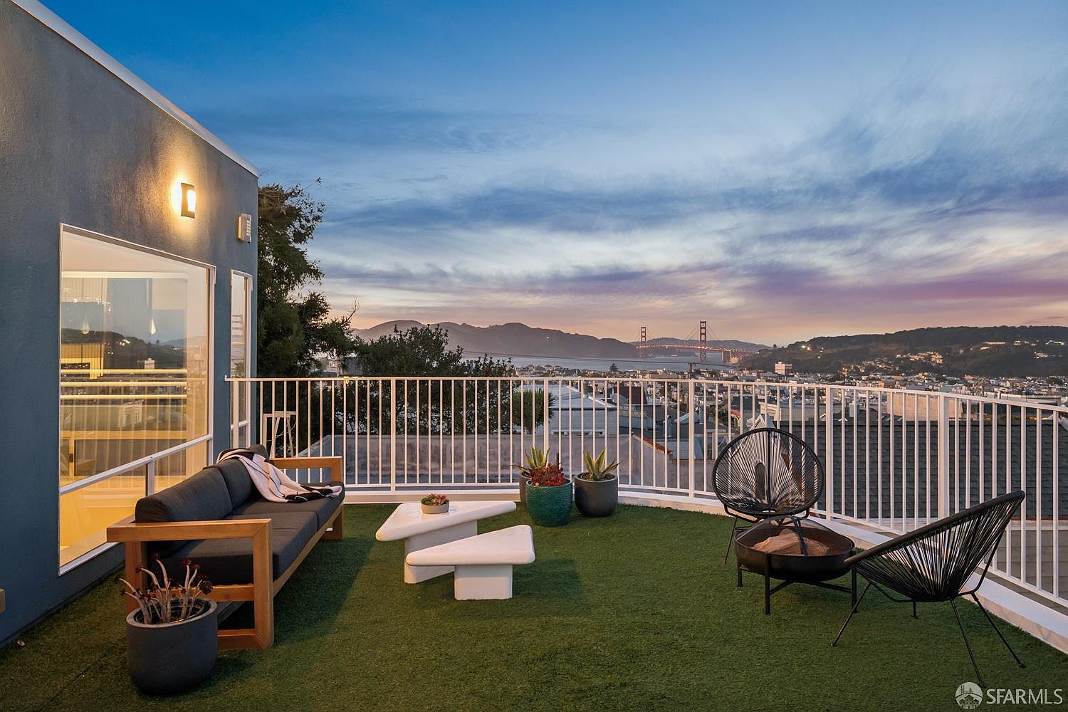 This elevated balcony features artificial turf flooring and a modern outdoor seating area, including a sofa and two wire-frame chairs arranged around a fire pit. The space is enclosed by a white railing and offers a panoramic, cinematic view of the city skyline and the Golden Gate Bridge at sunset. The scene is framed by a contemporary blue-toned exterior wall with integrated lighting, creating a sophisticated and inviting atmosphere for evening relaxation.