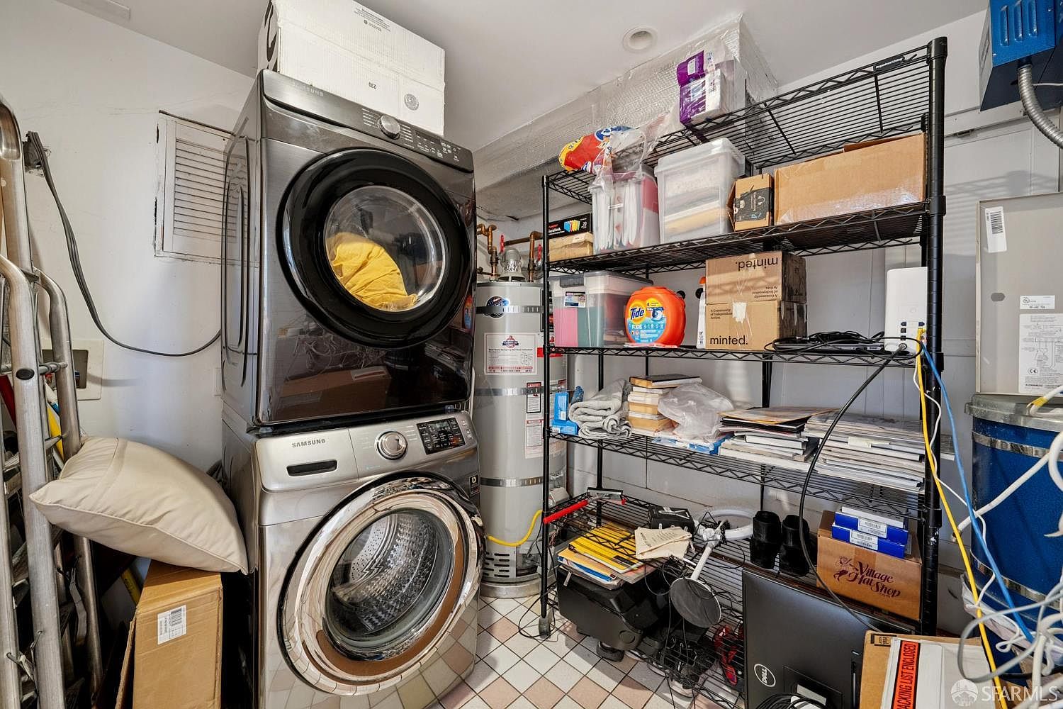 This utility space features a stacked Samsung washer and dryer unit positioned next to a tall water heater. A black metal wire shelving unit provides ample storage for household supplies, boxes, and miscellaneous items. The room has tiled flooring and appears to be a functional, utilitarian area within the home.