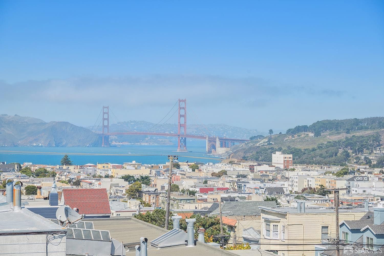 This elevated exterior view captures a panoramic cityscape of San Francisco, prominently featuring the iconic Golden Gate Bridge spanning the bay under a clear blue sky. The foreground showcases a dense urban residential neighborhood with various rooftops, chimneys, and utility lines, providing a sense of the property's location within the city. The perspective is from a high vantage point, likely a rooftop or upper-level deck, emphasizing the expansive and desirable views.