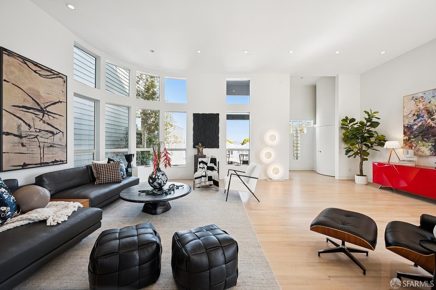 This expansive, light-filled living room features high ceilings and a curved wall of large windows that flood the space with natural light. The room is anchored by a dark leather sofa, a circular coffee table, and two black leather ottomans, all set atop a neutral area rug. A striking red console table adds a bold pop of color, while a tall indoor plant and modern art pieces contribute to the sophisticated, contemporary aesthetic.