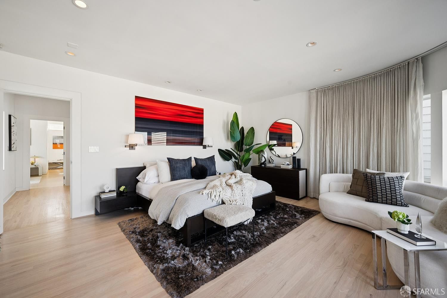 This spacious primary bedroom features a contemporary design with a dark wood bed frame, a plush textured rug, and a curved white sofa positioned near large windows draped in floor-to-ceiling curtains. The room is accented by a bold red and black abstract painting above the bed, a large round mirror, and a tall indoor plant that adds a touch of nature to the sophisticated space. The light wood flooring and neutral walls create a bright, airy atmosphere, while a doorway leads to an adjacent hallway, providing a sense of depth and flow.