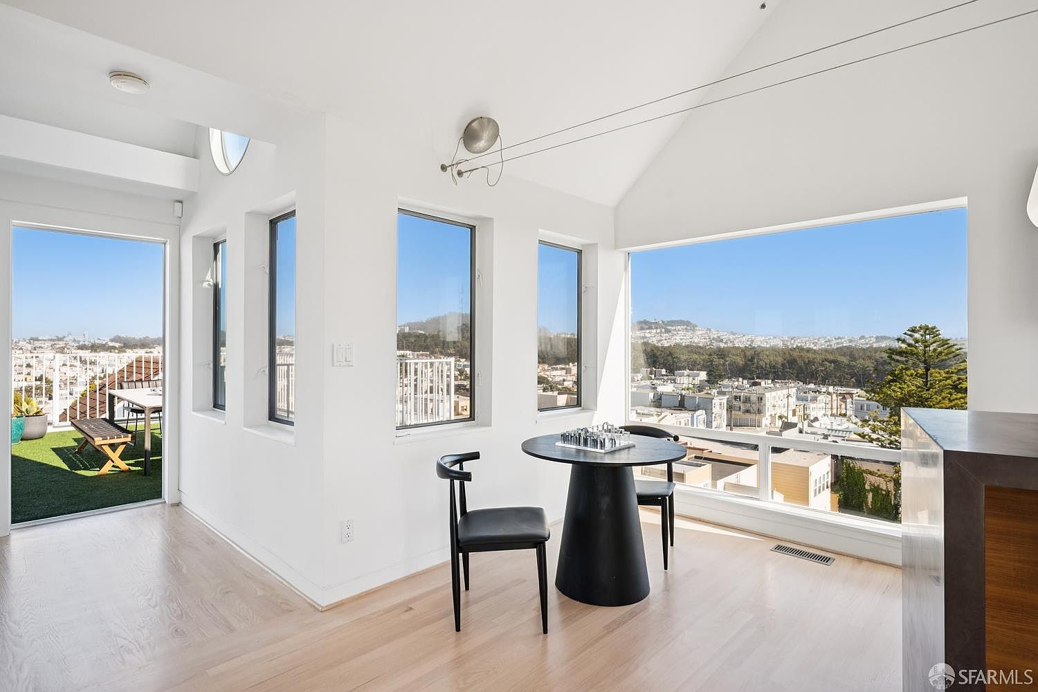This bright, modern dining area features a minimalist black circular table paired with two sleek black chairs, positioned to take advantage of the expansive city views through large windows. The room is characterized by its clean white walls, light wood flooring, and a vaulted ceiling that enhances the sense of openness. A glass door leads directly to an adjacent outdoor balcony, seamlessly blending indoor and outdoor living spaces.