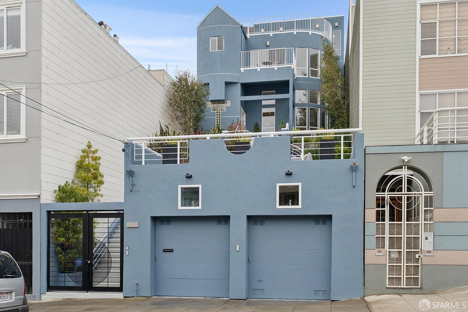 This front-facing view captures a multi-level blue residential building with a prominent two-car garage at street level. The facade features a large, elevated terrace with a white railing, multiple levels of balconies, and large windows that allow for significant natural light. The architectural style is contemporary, characterized by its stepped design and clean, modern lines.