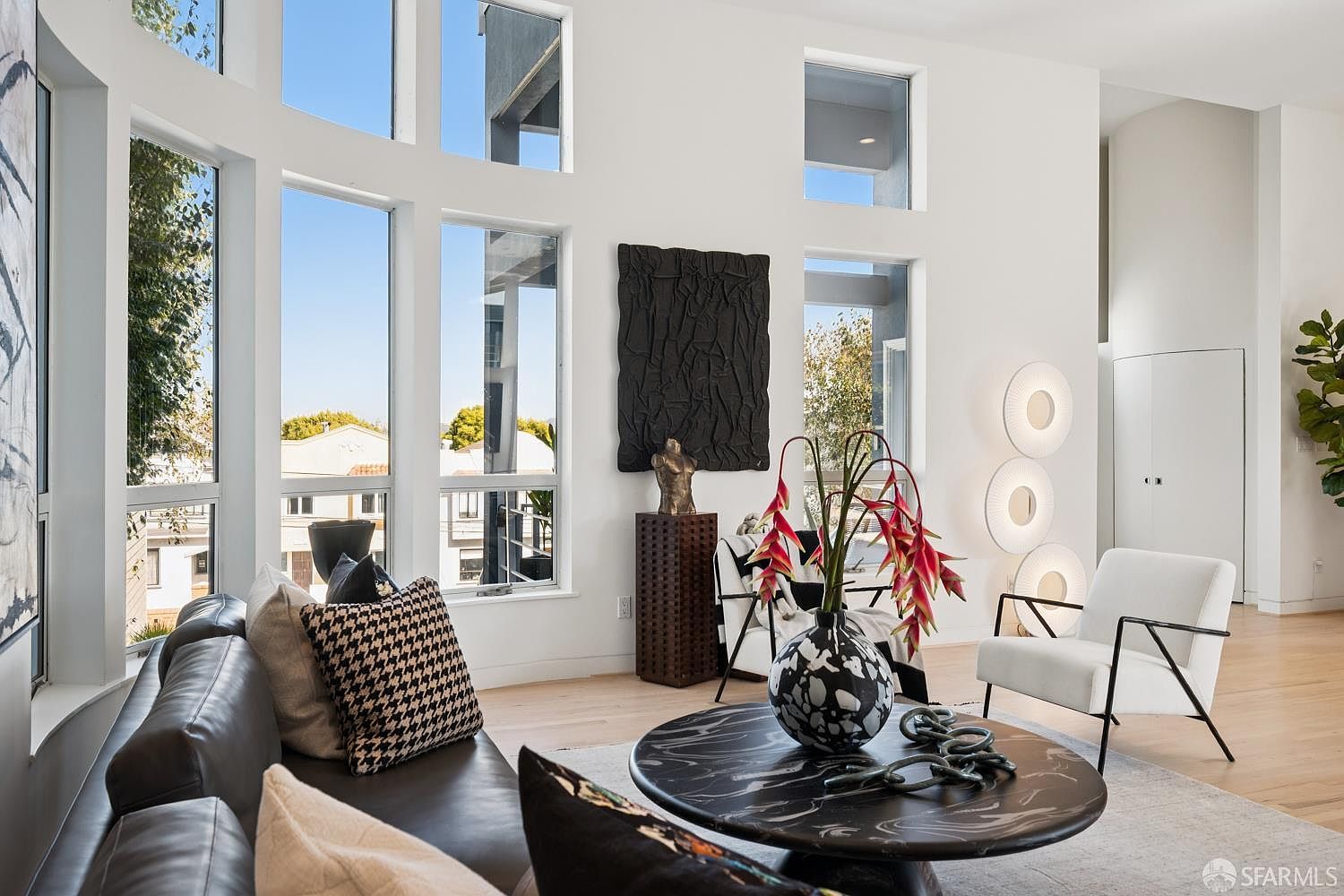 This bright and airy living room features a sophisticated, modern aesthetic with tall windows that flood the space with natural light. The room is anchored by a dark, circular stone coffee table and a plush leather sofa, complemented by a white accent chair and a unique sculptural floor lamp. A textured black wall hanging serves as a bold focal point, adding depth and artistic flair to the minimalist, high-ceilinged environment.