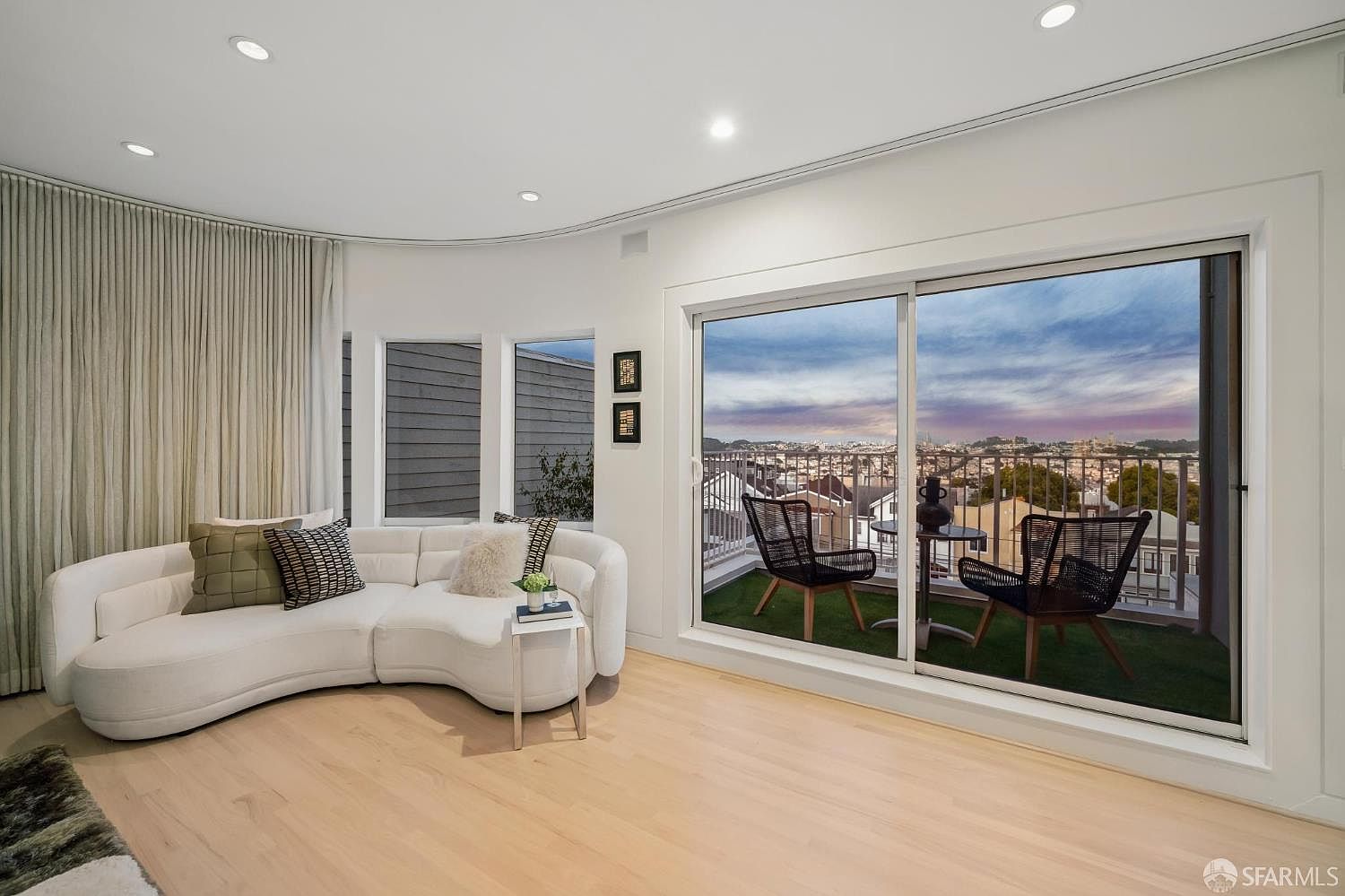 This bright and airy living room features a curved white sectional sofa positioned against a wall of tall windows draped in floor-length curtains. A sliding glass door opens onto a private balcony furnished with two chairs and a small table, offering an expansive view of the city skyline. The space is finished with light wood flooring and recessed ceiling lighting, creating a clean, modern, and inviting atmosphere.