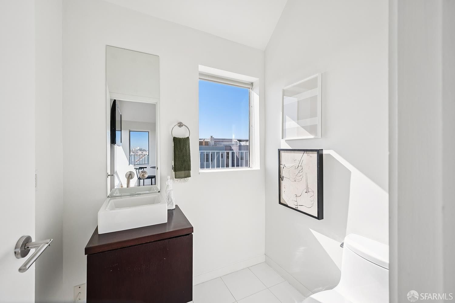 This minimalist powder room features a dark wood vanity topped with a white vessel sink and a tall, narrow mirror. A window above the vanity provides natural light and a view of the surrounding urban landscape, while two framed art pieces decorate the adjacent wall. The space is finished with clean white walls and light-colored tile flooring, creating a bright and modern aesthetic.