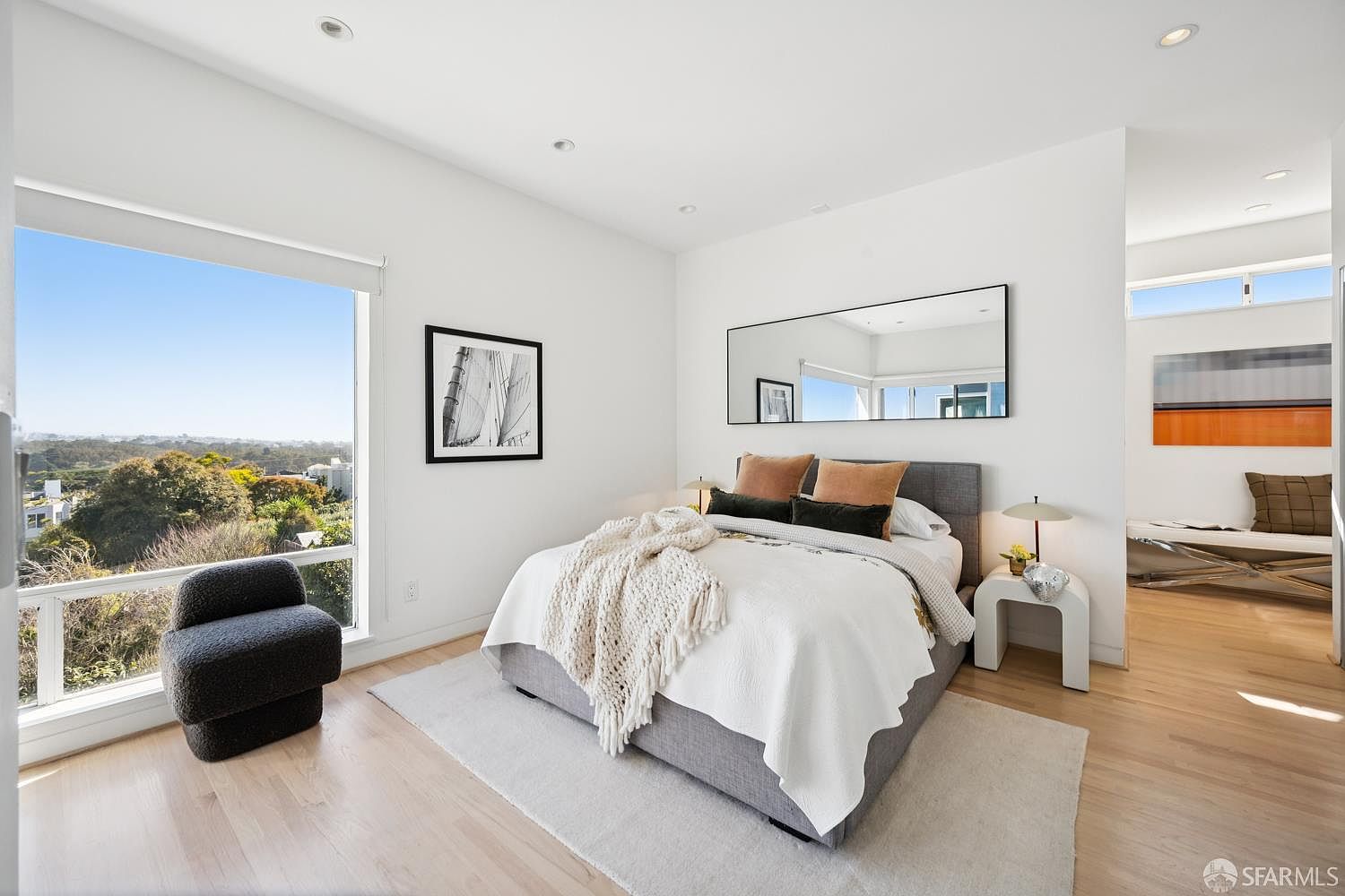This bright and airy primary bedroom features a large, grey upholstered bed centered against a wall with a wide, rectangular mirror. A dark grey accent chair sits near a large window that provides an expansive view of the surrounding landscape. The room is finished with light wood flooring, a neutral area rug, and a minimalist side table, creating a serene and contemporary atmosphere.