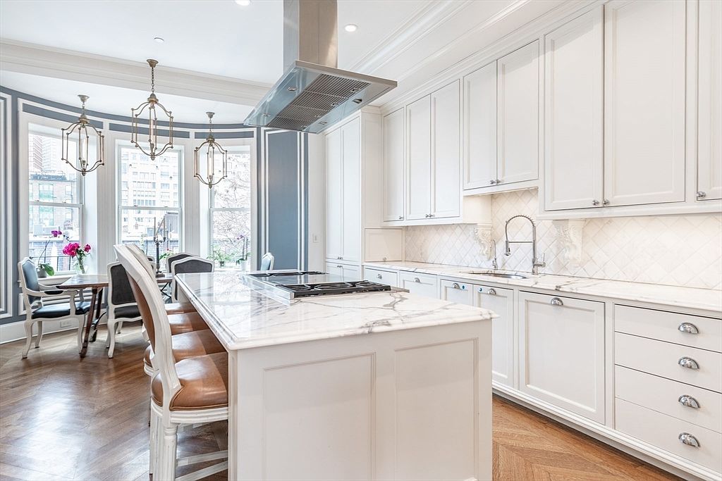 This elegant, bright kitchen features white cabinetry, a large central island with a stone countertop, and a built-in gas cooktop. A dining area with a table and chairs is positioned near a bay window, creating a seamless transition between cooking and eating spaces. The room is accented by classic pendant lighting, a herringbone-patterned wood floor, and a neutral color palette that enhances the sophisticated, airy atmosphere.