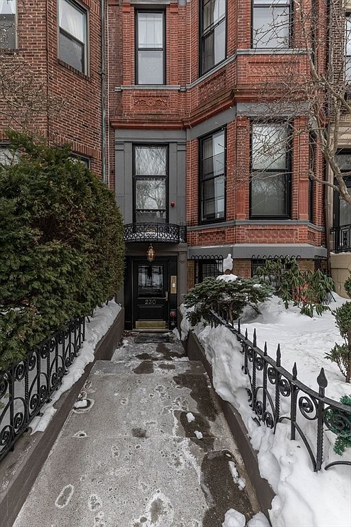This image captures the front entryway of a classic brick townhouse, accessed by a stone walkway flanked by decorative black iron fencing. The entrance features a dark-painted door with a small balcony above, set against a backdrop of traditional masonry and tall, multi-pane windows. The scene is framed by snow-covered landscaping, suggesting a cold, winter setting that highlights the home's historic urban character.