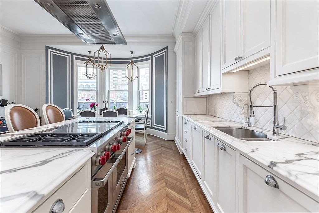 This elegant kitchen features a central island with a gas range and a professional-grade hood, complemented by white cabinetry and stone countertops. The space is anchored by rich, herringbone-patterned wood flooring and a curved bay window area that serves as a bright breakfast nook. The perspective is a straight-on shot looking down the galley-style layout, highlighting the sophisticated contrast between the light cabinetry and the dark, paneled wall accents.