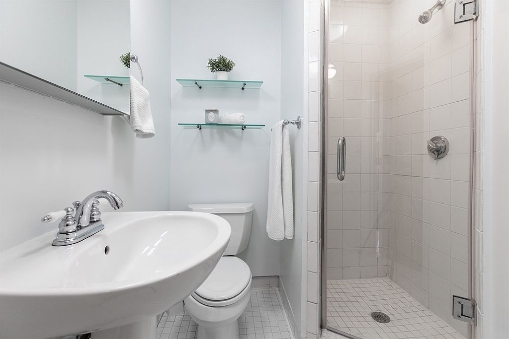 This compact, functional bathroom features a white pedestal sink, a standard toilet, and a glass-enclosed shower stall with white tiled walls. Two glass shelves mounted on the wall provide storage, while a mirror above the sink enhances the sense of space. The room is finished with light-colored walls and white floor tiles, creating a clean and bright aesthetic.