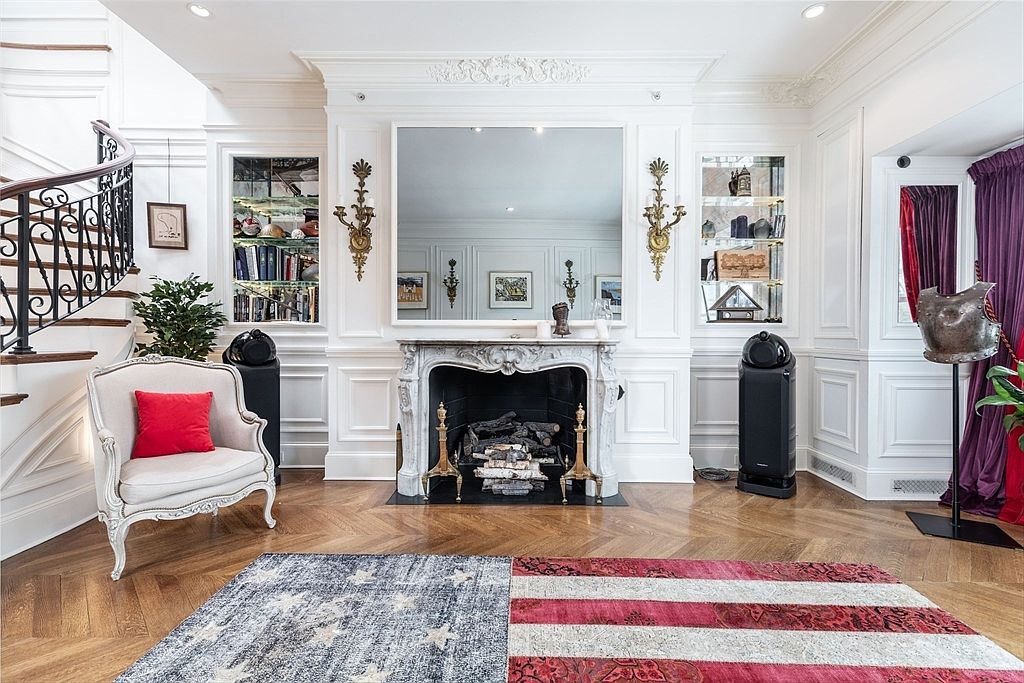 This elegant living room features a grand, ornate stone fireplace surround centered between two built-in glass-front display cabinets. A classic staircase with a decorative iron railing curves along the left side, while a large, patterned area rug featuring an American flag motif anchors the space. The room is finished with intricate wall molding, polished herringbone wood floors, and sophisticated wall sconces, creating a refined and stately atmosphere.