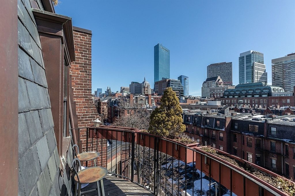 This elevated balcony offers a panoramic view of the city skyline, featuring a prominent skyscraper and historic brick row houses. The space is furnished with two small metal bistro chairs and a table, set against a backdrop of slate-tiled siding and a brick chimney. The perspective captures a clear, sunny day, highlighting the urban setting and the private outdoor living area.