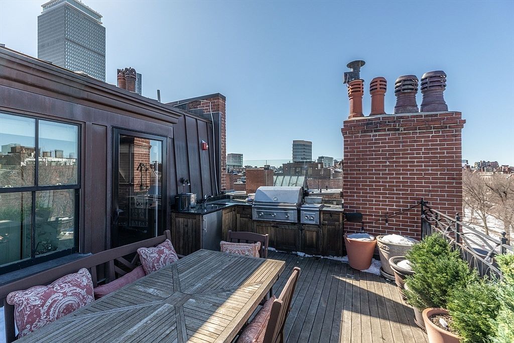 This rooftop deck offers an expansive outdoor living space featuring a wooden dining set and a built-in outdoor kitchen with a stainless steel grill. The area is framed by a brick chimney structure with multiple flues and provides panoramic views of the surrounding city skyline. The perspective is elevated, capturing the blend of urban architecture and private outdoor relaxation space.