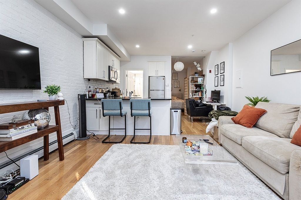 This open-concept living space features a bright, modern aesthetic with a white-painted brick wall and light wood flooring. The room includes a comfortable sofa with orange accent pillows, a clear coffee table, and a breakfast bar with two stools that connects to the kitchen area. A television is mounted on the brick wall above a wooden console, creating a cozy and functional layout that flows into a secondary living or office space in the background.