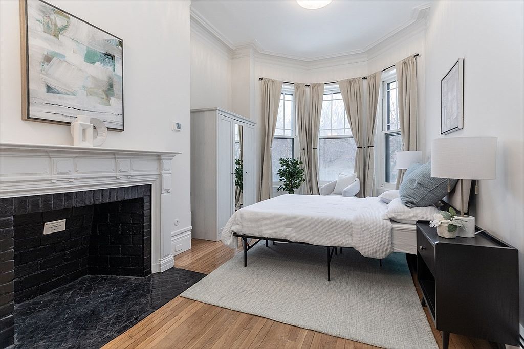 This bright and airy bedroom features a prominent black brick fireplace with a white mantel, serving as a striking focal point. The room is furnished with a bed centered on a light-colored area rug, positioned in front of a bay window dressed with neutral curtains. A tall white wardrobe and a dark bedside table provide functional storage, while the light wood flooring and white walls create a clean, classic aesthetic.